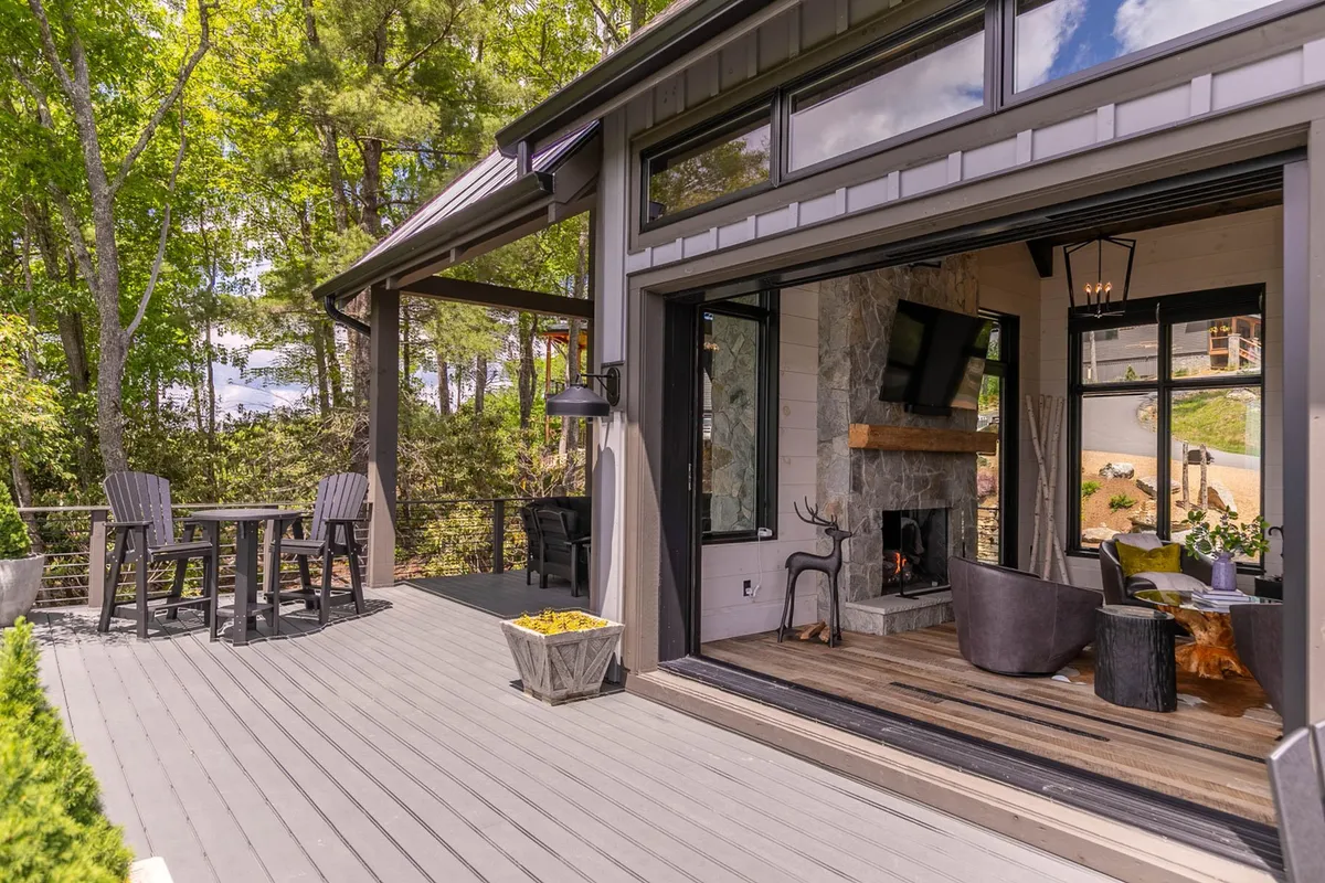 A tranquil outdoor seating area features a stone fireplace surrounded by comfortable chairs on a wooden deck. Large windows connect the patio to the house, and lush greenery provides a peaceful backdrop. The space is set for social gatherings, relaxing by the fire, and enjoying the mountain air.