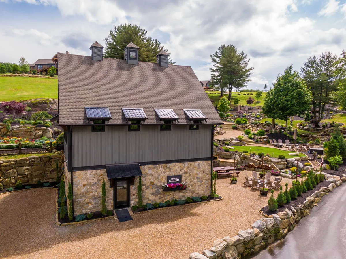 This image displays the rustic stone façade of the building, surrounded by a landscaped garden with an outdoor seating area, and a gravel driveway leading up to the entrance. The setting is open and inviting, suggesting a communal space where outdoor activities and gatherings can take place.