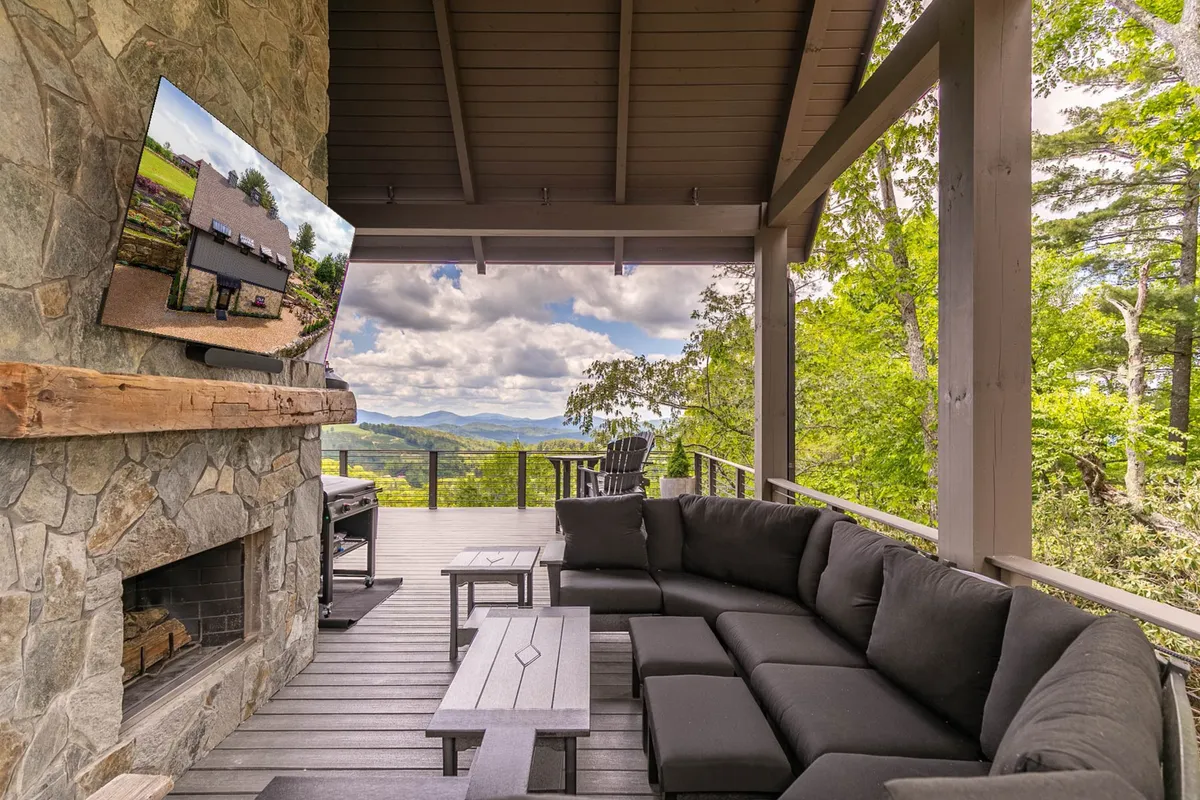 This scene features an expansive outdoor lounge area with a stone fireplace, a large sectional couch, a TV mounted above the fireplace, and sweeping mountain views. The atmosphere is casual and comfortable, ideal for lounging and group gatherings.
