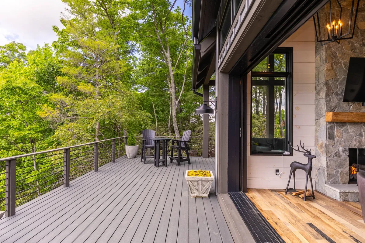 A spacious wooden deck surrounded by trees, featuring an inviting outdoor seating area and a cozy fireplace. The setting opens up to tranquil nature views, suggesting a perfect spot for gatherings or relaxation in the fresh mountain air.