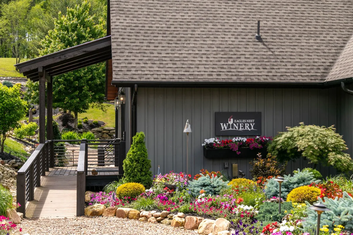 This exterior image features the welcoming entrance of the Eagles Nest Winery, highlighted by a prominent winery sign, beautiful flower beds, a wooden walkway, and lush greenery. The vibrant landscaping and natural stone pathway create an inviting, upscale ambiance that aligns with the experience of strolling to a nearby winery for hand-selected wines.