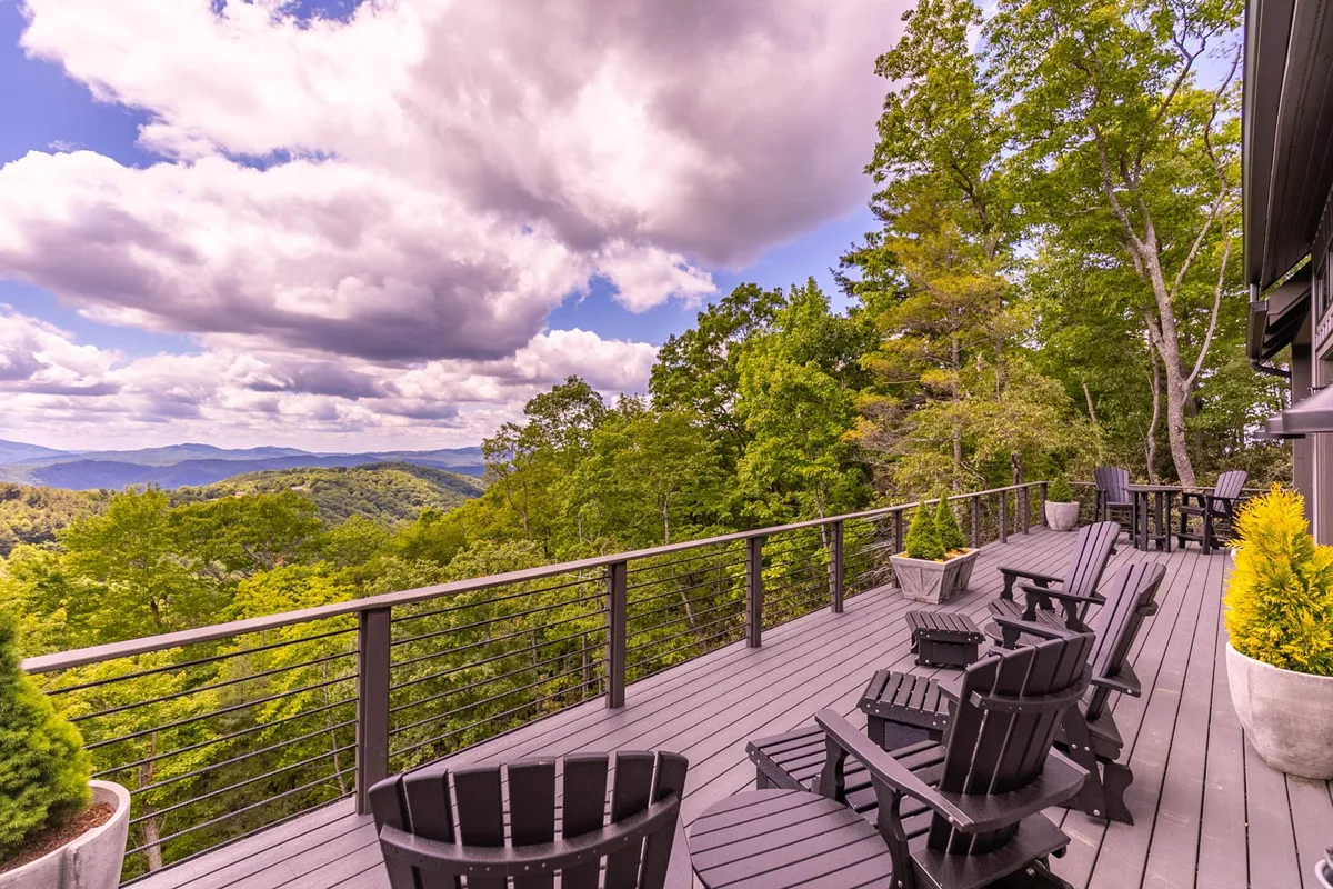 A spacious deck with adirondack chairs overlooks a sweeping mountain vista and lush greenery. The sky is partly cloudy, suggesting changing seasons. The inviting setup encourages outdoor relaxation, socializing, and taking in the natural beauty from sunrise to sunset.