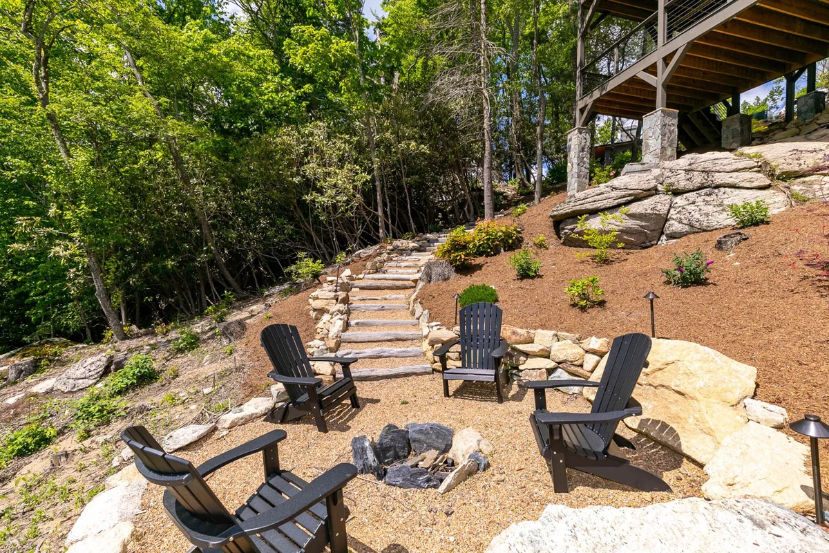 The image depicts a welcoming fire pit area surrounded by black Adirondack chairs, set amidst landscaped gardens and stone steps. The mood is cozy and communal, inviting guests to share stories and s’mores under the open sky.
