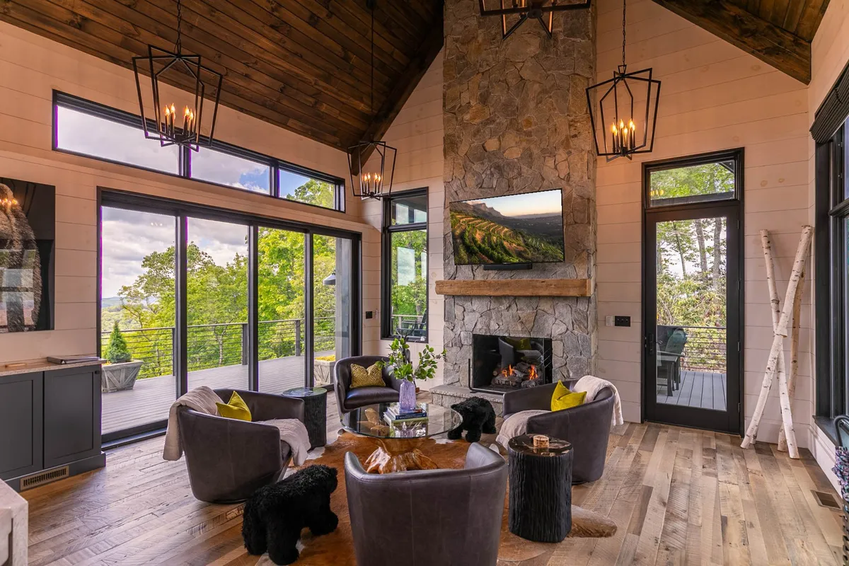 A cozy living area anchored by a stone fireplace. Plush seating faces large windows that flood the room with natural light and provide captivating mountain scenery. The inviting mood is enhanced by a wooden coffee table and soft decor accents.