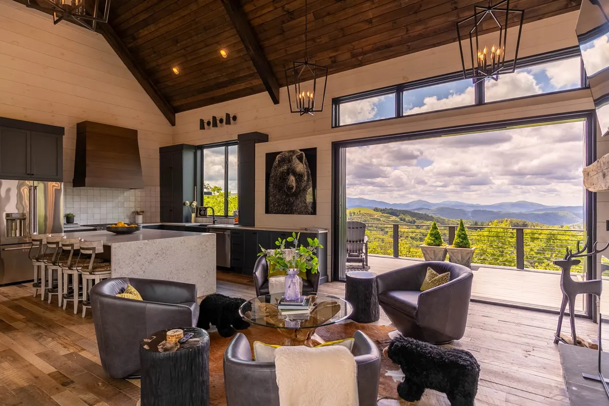 A modern kitchen with a large granite island, sleek appliances, and panoramic windows framing mountain vistas. The space includes stylish furniture and eye-catching black bear artwork, enhancing both the culinary and social aspects of the room.