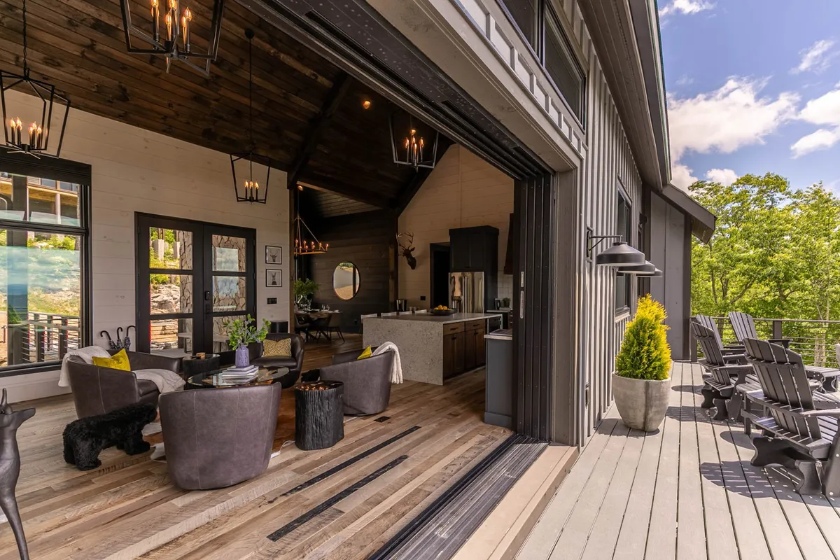 An expansive view of the open-concept living area displaying a wooden ceiling, modern furniture, and floor-to-ceiling windows that open to a surrounding deck. The scene captures the seamless connection between indoors and the outdoors, with visible sunset hues through the windows and an inviting atmosphere perfect for gatherings.