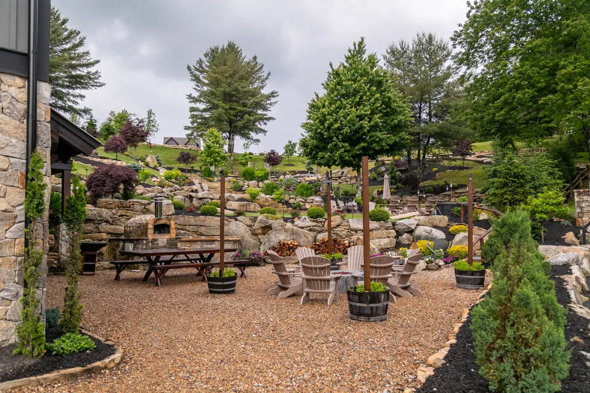 A thoughtfully designed outdoor space with stone seating areas, a large dining table, lush greenery, and dramatic rock formations. The communal table and natural setting invite guests to dine or celebrate together outdoors.