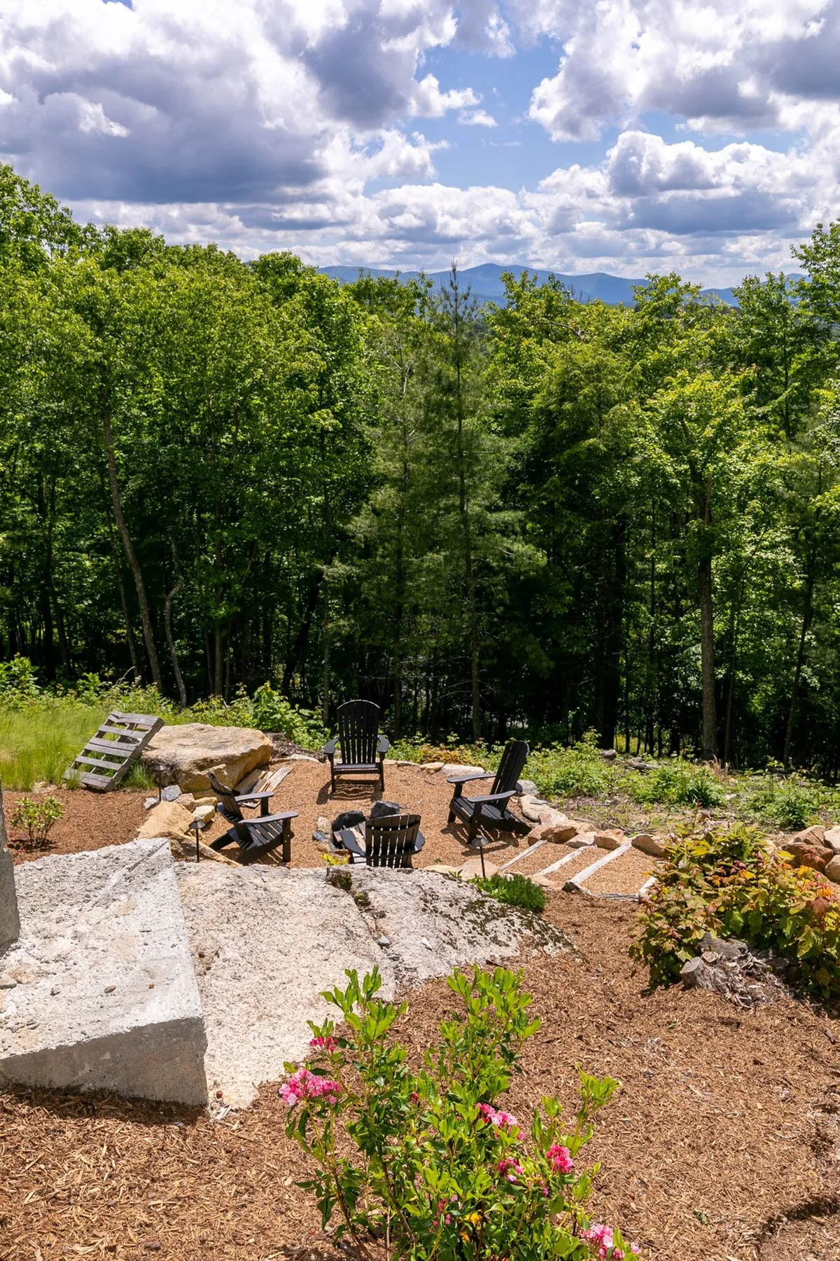 A cozy fire pit area surrounded by black Adirondack chairs and flowering plants creates an idyllic spot for evening gatherings. The space is bordered by a natural stone pathway and lush greenery, highlighting opportunities for relaxation and storytelling after mountain activities.